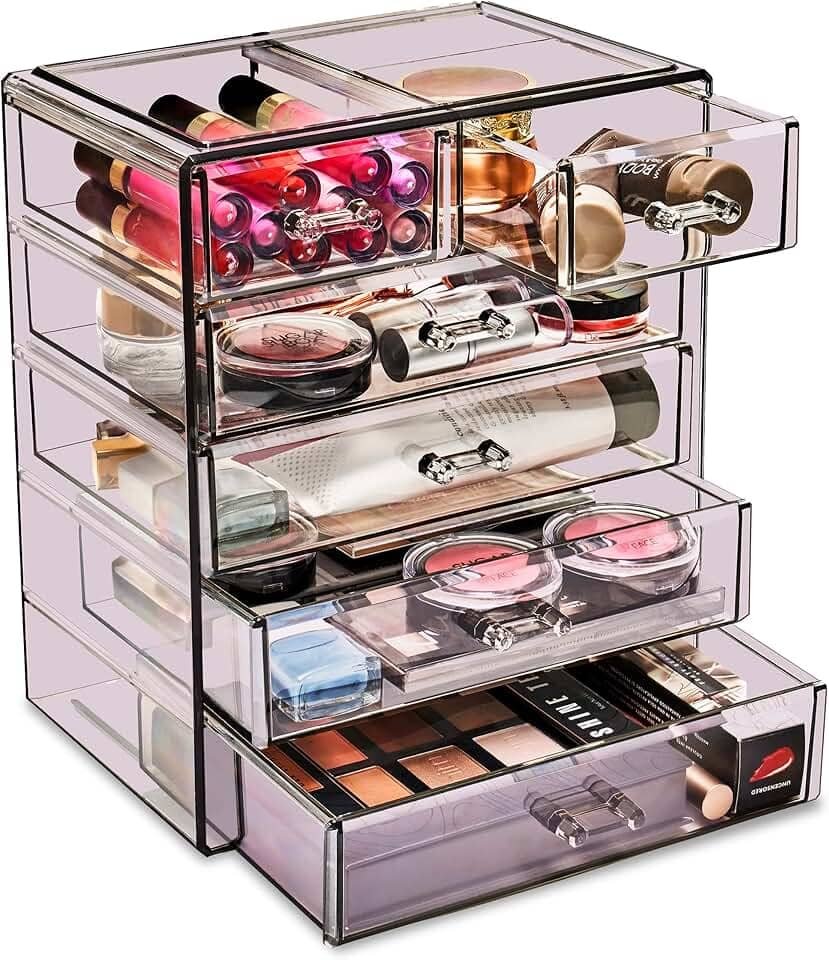 A clear Sorbus Acrylic Makeup Organizer with six drawers and multiple compartments neatly displaying lipsticks, brushes, and jewelry on a vanity countertop.